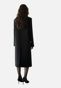 Lange zwarte jas met een rechte, op maat gemaakte ontwerp, voorzien van een naad in het midden van de rug en lange mouwen. Draag het over doorschijnende zwarte kousen en hakken.