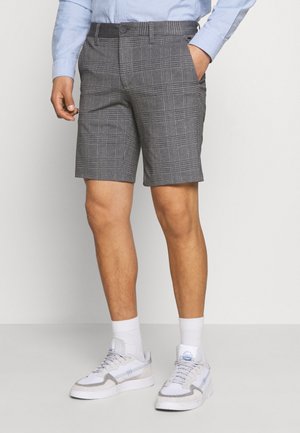 Mann trägt graue karierte Shorts, weiße Socken, weiße Sneaker und ein hellblaues langärmeliges Hemd, eine Hand in der Tasche.
