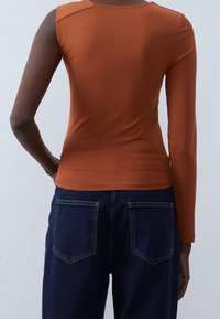 Langärmliges, tailliertes Oberteil in verbranntem Orange, aus glattem Stoff. Kombiniert mit hoch taillierten, dunklen Jeans aus Denim mit zwei Gesäßtaschen.