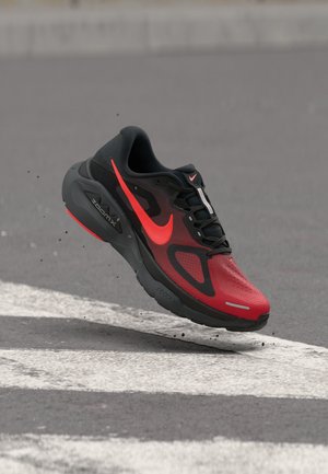 Black and red Nike ZoomX running shoe mid-air above asphalt with white road markings and scattered small debris.
