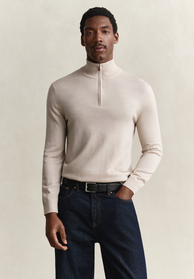 Homme portant un pull beige à demi-fermeture éclair, jean foncé et ceinture noire, debout avec une main dans la poche sur fond uni.