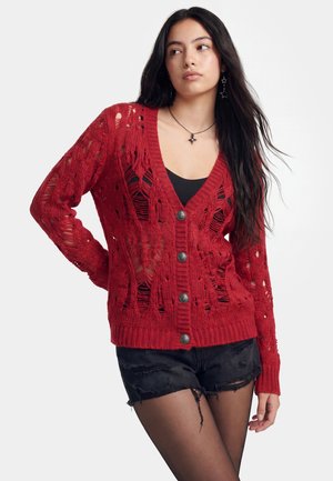 Ung kvinde med langt mørkt hår iført en rød knap-på-strikket cardigan, sort tanktop, sorte slidte shorts og smykker formet som stjerner.