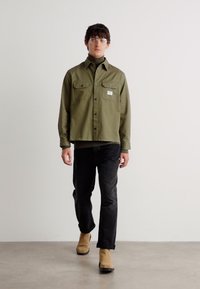 Olive green button-up shirt with two chest pockets, worn over a black turtleneck, paired with dark jeans and tan ankle boots.