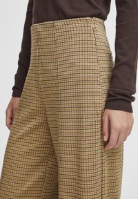 Pantalon à carreaux beige et noir, doté d'une taille haute, de jambes larges et d'une texture subtile, agrémenté d'une fermeture éclair latérale discrète.
