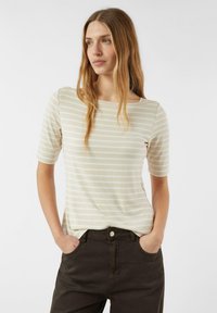 Bawełniany t-shirt w paski w kolorze kremowym i jasnobeżowym, z dekoltem typu łódka i krótkimi rękawami, zestawiony z ciemnobrązowymi spodniami.