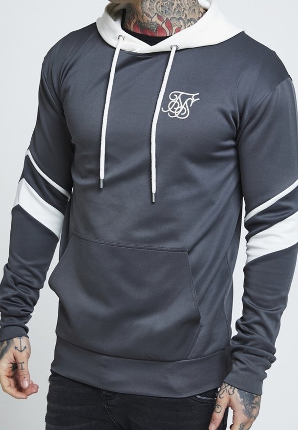 Mężczyzna w szarym kapturze z białym kapturem, białymi sznurkami, białymi paskami na rękawach i logiem na klatce piersiowej, z tatuażami na szyi i rękach.