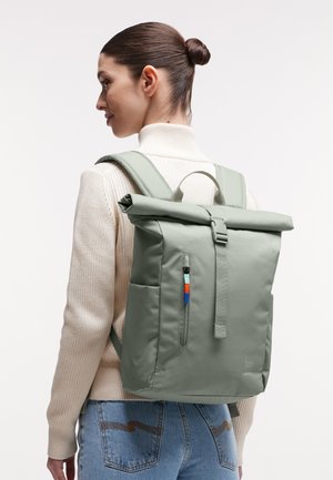 Femme aux cheveux attachés en chignon, portant un col roulé beige et un jean bleu, portant un sac à dos vert clair à fermeture roulante avec une poche zippée à l'avant.
