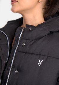 Giubbotto imbottito nero con collo alto, zip e bottoni automatici. Presenta un logo bianco ricamato sul petto e un tessuto testurizzato.