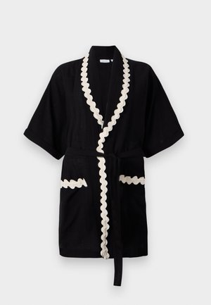 Túnica negra de mangas curtas, com acabamento creme em ondas ao longo do colarinho e dos bolsos, apresentando um cinto a condizer. Tecido texturizado.