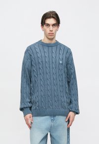 Μπλε πουλόβερ πλεκτό με στρογγυλή λαιμόκοψη και ριμπ μανσέτες. Διαθέτει έντονες στριφτές λεπτομέρειες και μικρό λογότυπο στο στήθος.