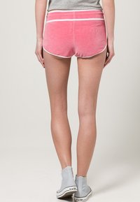 Shorts en velours rose avec des passepoils blancs, arborant un design taille basse. Portés avec des bottines grises à la cheville. La vue de dos met en valeur la texture douce.