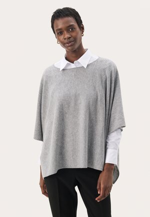 Poncho grigio chiaro in morbido tessuto, con un design rilassato e maniche ampie, indossato sopra una camicia bianca con colletto e pantaloni neri.
