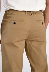 Beige Baumwollhosen mit einem elastischen Bund, ausgestattet mit zwei Gesäßtaschen mit Knopfverschluss und einem glatten, strukturierten Stoff.