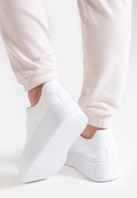 Białe sneakersy na platformie o gładkiej teksturze i zaokrąglonym kształcie, zestawione z jasnoróżowymi dresami z haftem "Easy to lab" na mankiecie.