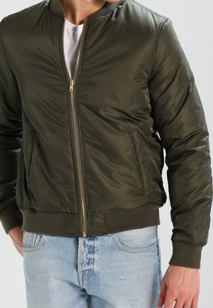 Homme portant une veste bomber zippée vert olive sur un t-shirt blanc et un jean bleu clair, debout devant un fond uni.