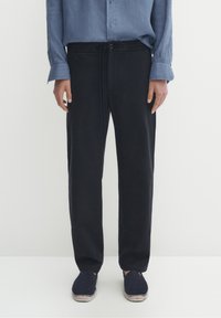 Donkerblauwe elastische broek met een koord in de taille, gecombineerd met een lichtblauw overhemd met knopen, en marineblauwe espadrilles met een gestructureerde zool.
