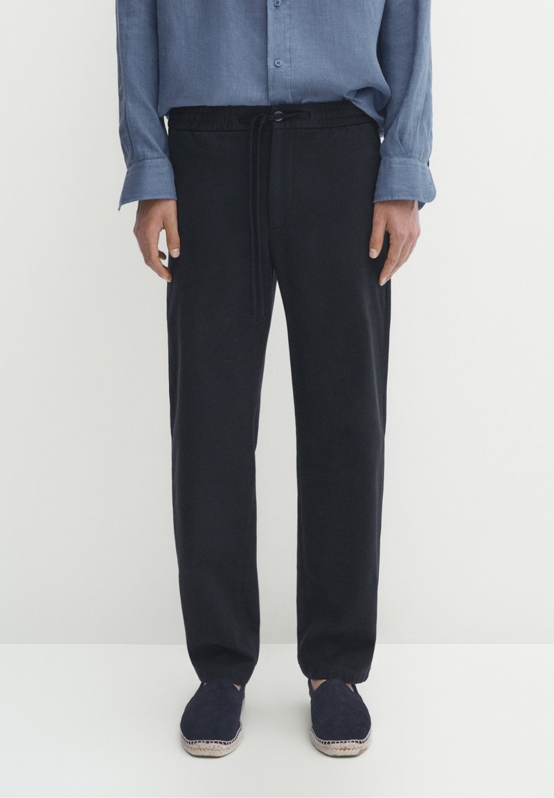 Donkerblauwe elastische broek met een koord in de taille, gecombineerd met een lichtblauw overhemd met knopen, en marineblauwe espadrilles met een gestructureerde zool.