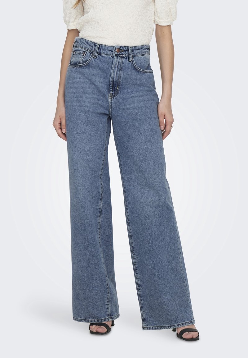 Femme portant un jean bleu taille haute à jambes larges, associé à un haut blanc texturé à manches courtes et des talons ouverts noirs.