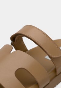 Sandales couleur beige avec larges brides, présentant un design découpé. Texture lisse et mate avec des détails de surpiqûres et un léger talon.