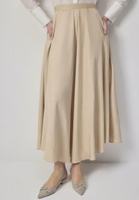 Woman wearing a beige, flowy midi skirt with hands in pockets, paired with pointed beige flats decorated with rhinestone bows.