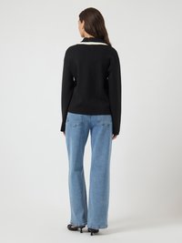 Kvinne som vender bort fra kameraet, iført svart langermet genser med hvit kragedetalj, lyseblå rette jeans og svarte kitten-heeled sko.