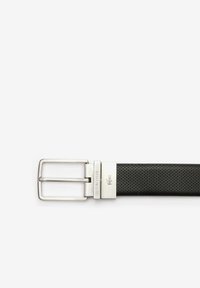Cinturón de cuero perforado negro con hebilla de metal plateado. Presenta el logo grabado "LACOSTE" en la hebilla. Diseño rectangular clásico.