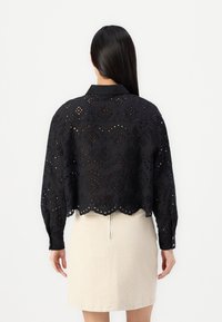 Black, cropped blouse with intricate cut-out patterns, long sleeves, and scalloped hem, paired with a light-colored skirt.