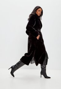 Zwarte nepbontjas met een ceintuur in de taille, lange mouwen en zijsplitten, gecombineerd met zwarte kniehoge stiletto laarzen.