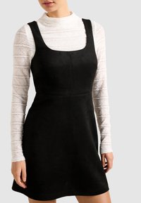 Femme portant une robe noire sans manches sur un top à manches longues en dentelle blanche à col montant, se tenant devant un fond clair uniforme.
