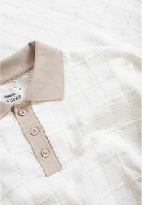 Polo shirt en tricot blanc avec un col et une patte beige à côtes, comportant trois boutons marron clair et un motif en losanges texturé.