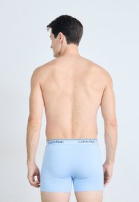 Man staat met zijn rug naar de kijker, gekleed in lichtblauwe Calvin Klein boxershorts, met een effen witte achtergrond.