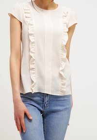 Světle broskvová blůza s krátkými rukávy, volantové detaily na přední straně, strukturovaná látka, zkombinovaná s modrými džíny.