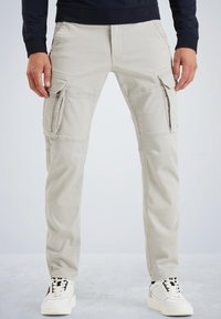 Light beige cargo pants featuring multiple pockets, zip closures, and a straight leg design. Paired with white sneakers for contrast.