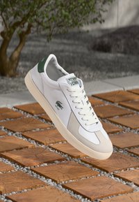 White leather sneaker with green accents, featuring flat laces, a cushioned sole, and a textured toe cap. Lacoste logo displayed prominently.