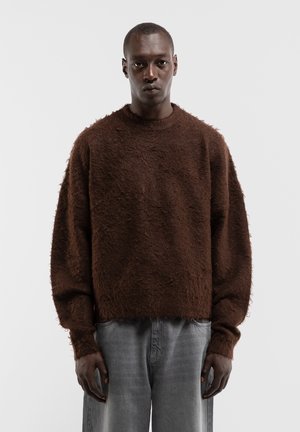 Mann trägt einen lockeren, strukturierten braunen Pullover und hellgraue Jeans und steht vor einem schlichten hellen Hintergrund.