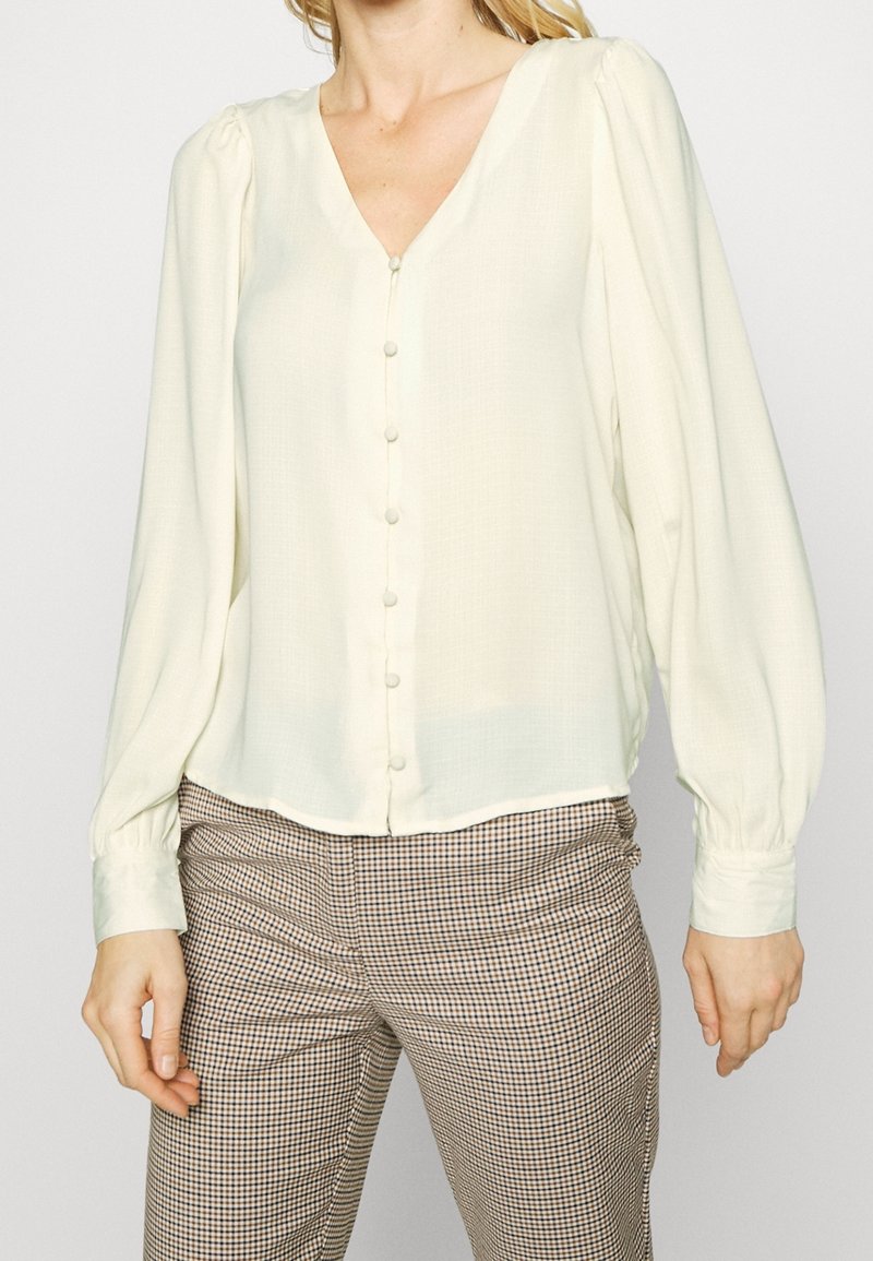 Femme portant un chemisier crème à manches longues avec des boutons cachés et des épaules bouffantes, assorti à un pantalon à carreaux marron et blanc.