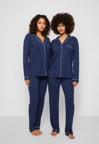 Mörkblå pyjamasset med långa ärmar, knäppning fram och vita pipingar. Mjuk tyg, avslappnad passform, kompletterad med matchande byxor.