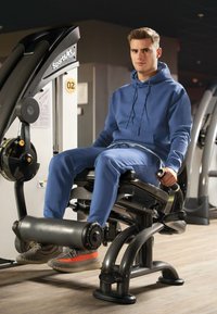 Completo di felpa blu e pantaloni da tuta indossato da una persona seduta su una macchina da fitness nera con accenti argento in un ambiente da gym.