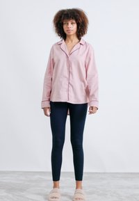 Ljusrosa skjorta med mörka detaljer, kombinerad med marinblå leggings och beige tofflor. Modellen står på en ljusgrå matta mot en vit bakgrund.