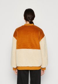 Parte superior de sarga color óxido con textura cremosa y lanosa en la parte inferior. Presenta un corte relajado y cuello redondo; puños y dobladillo acanalados.