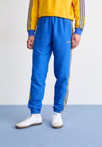 Pantalones de chándal azules con franjas laterales amarillas, cinturilla elástica y puños, combinados con zapatillas blancas. El material parece brillante y suave.