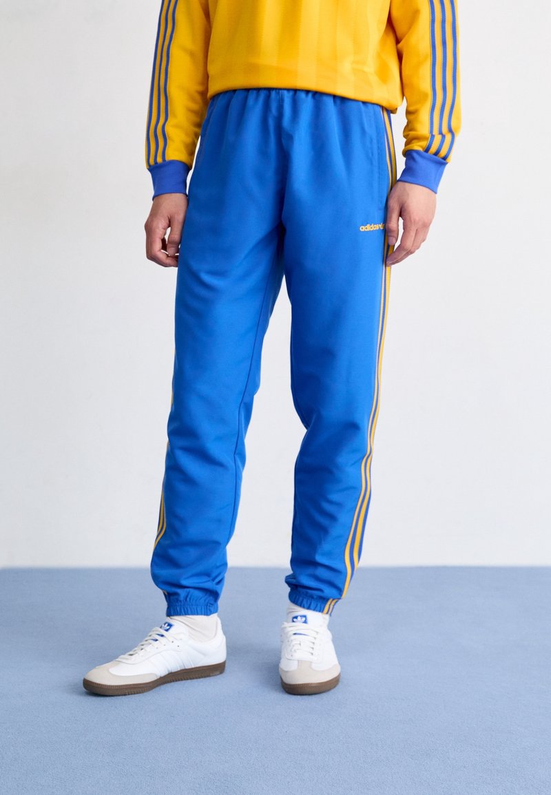 Pantalones de chándal azules con franjas laterales amarillas, cinturilla elástica y puños, combinados con zapatillas blancas. El material parece brillante y suave.