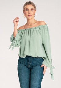 Jasnozielona bluzka hiszpanka z marszczonym dekoltem i wiązanymi rękawami, wykonana z miękkiego materiału, zestawiona z ciemnymi dżinsami z wysokim stanem.