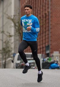 Blauer Hoodie mit fettem weißen Text, schwarze Leggings und schwarze Sportschuhe. Läufer auf einer Stadtstraße, trägt weiße Socken und hat einen konzentrierten Ausdruck.