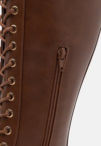 Braune Lederstiefel mit einem seitlichen Reißverschluss und goldfarbenen Ösen mit Schnürsenkeln. Die Oberfläche hat eine glatte Textur und eine enganliegende Form.