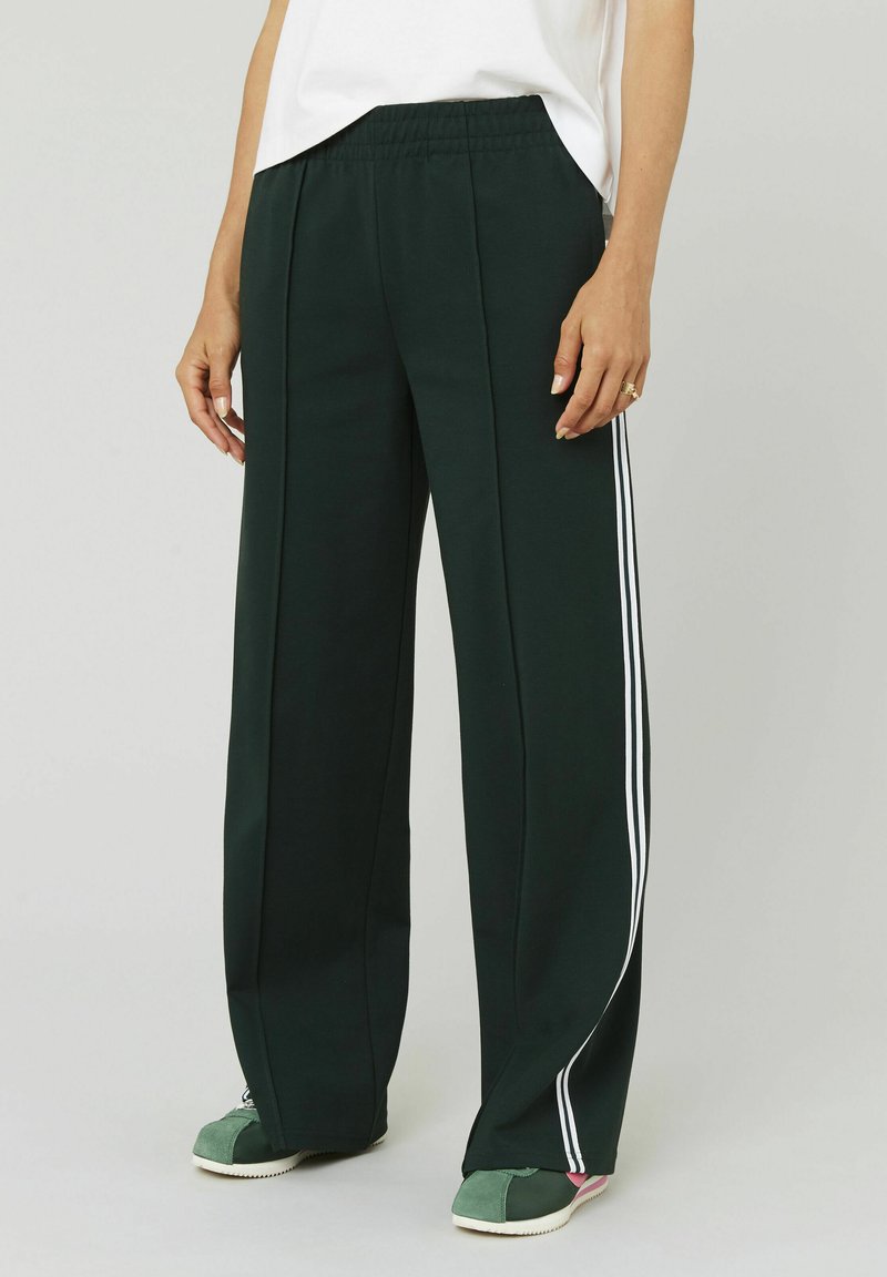 Pantalons à jambes larges vert foncé avec une texture douce. Présentent des bandes blanches sur les côtés et une taille élastique. Assortis à des baskets vertes.