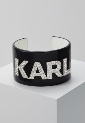 Bracelet manchette noir avec grandes lettres argentées « KARL » ornées de strass clairs, présenté sur un socle blanc sur fond gris.