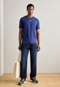 Blå bomulls t-skjorte med korte ermer, kombinert med mørk denimjeans og flerfargede joggesko. Individet holder en lys totebag.