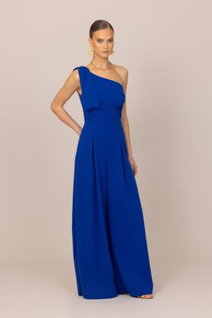 Kobieta z gładko zaczesanymi włosami, nosząca złote kolczyki i naszyjnik, pozująca w królewskim niebieskim kombinezonie na jedno ramię z szerokimi nogawkami na beżowym tle.