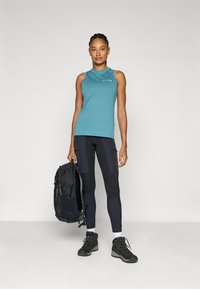Tiefer, ärmelloser Oberteil in Petrol, schwarze Leggings mit Taschen, schwarze Wanderschuhe und ein schwarzer Rucksack. Einfaches Design, leichtes Gewebe und strapazierfähige Texturen.